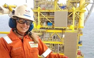 Woman wearing orange boiler suit and white helmet with yellow oil rig behind her